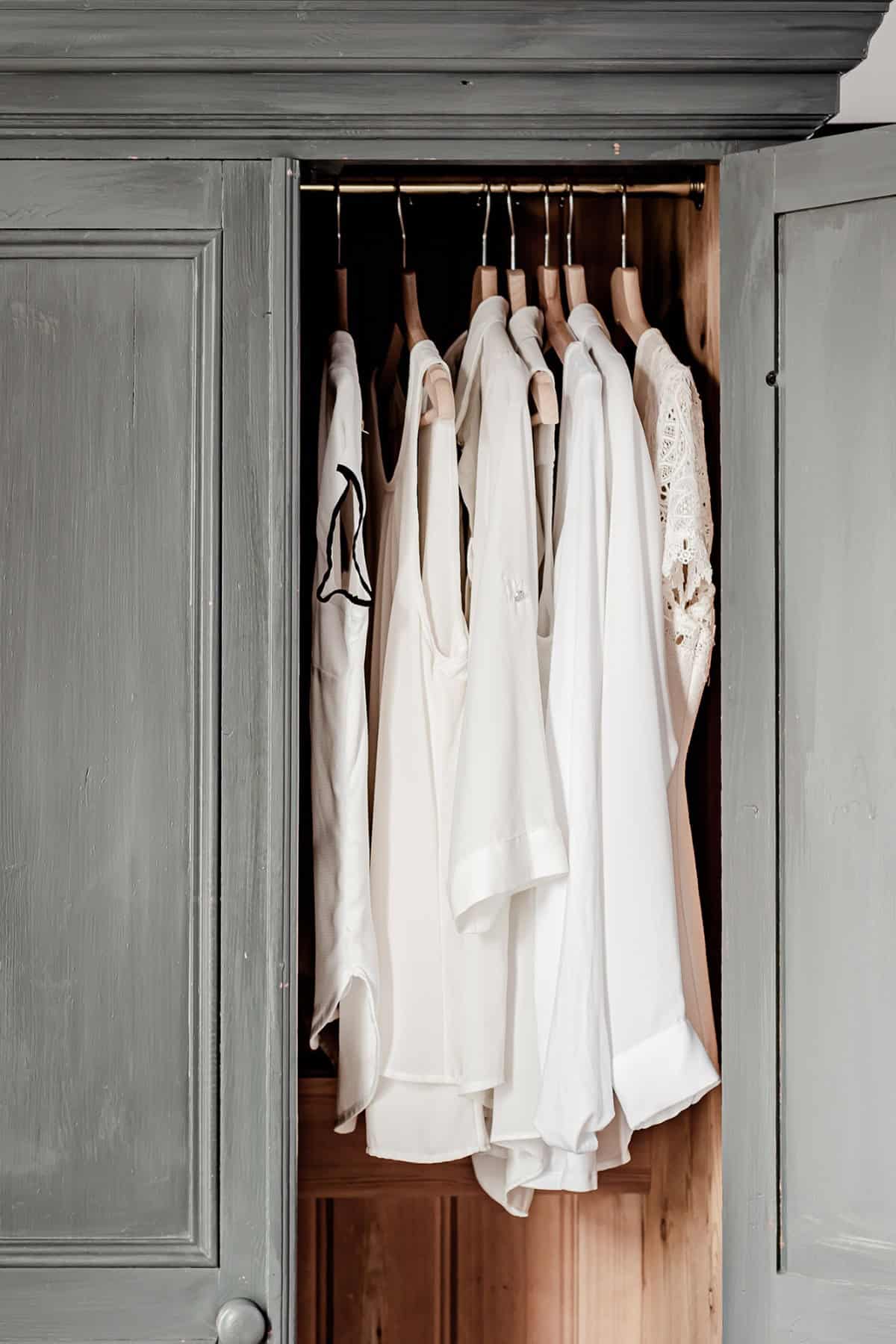 A partially open, rustic grey wardrobe reveals several white clothing items hanging neatly inside on wooden hangers. The clothing includes various styles of tops and blouses, contributing to an organized and minimalist aesthetic.