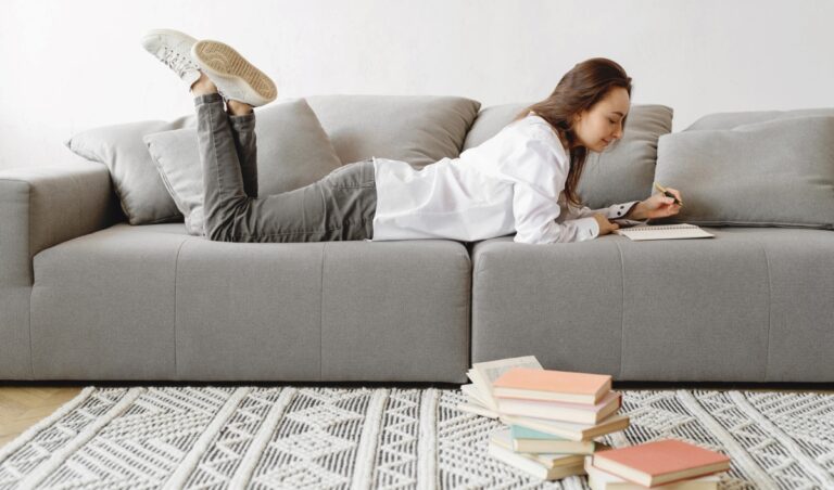 A woman lounges on a sleek gray sectional sofa, writing in a notebook while surrounded by a scattered stack of books on a patterned rug, capturing a cozy moment of messy minimalism. Her casual outfit and the neutral-toned room reflect a lived-in yet uncluttered aesthetic.