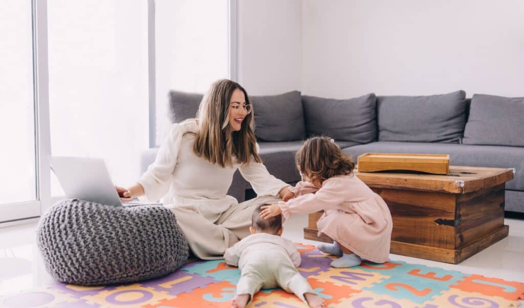 A mother sits on a colorful alphabet foam mat with her laptop nearby, smiling as her toddler gently pats a crawling baby. The cozy living room features a gray sectional sofa and a rustic wooden chest, blending work-from-home life with playful parenting moments.