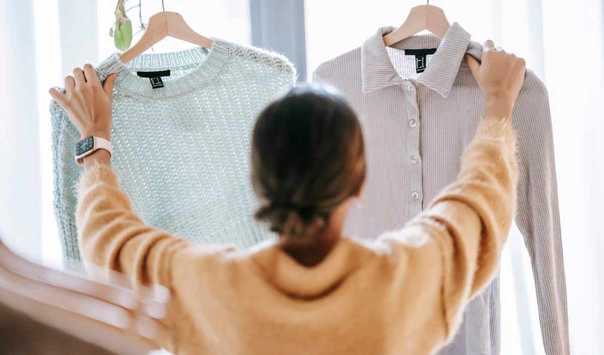 Woman seen from behind holding up two sweaters on hangers—one light blue and loosely knit, the other beige with buttons and a collar—while deciding between them, illustrating a minimalist or “one in, one out” wardrobe choice.