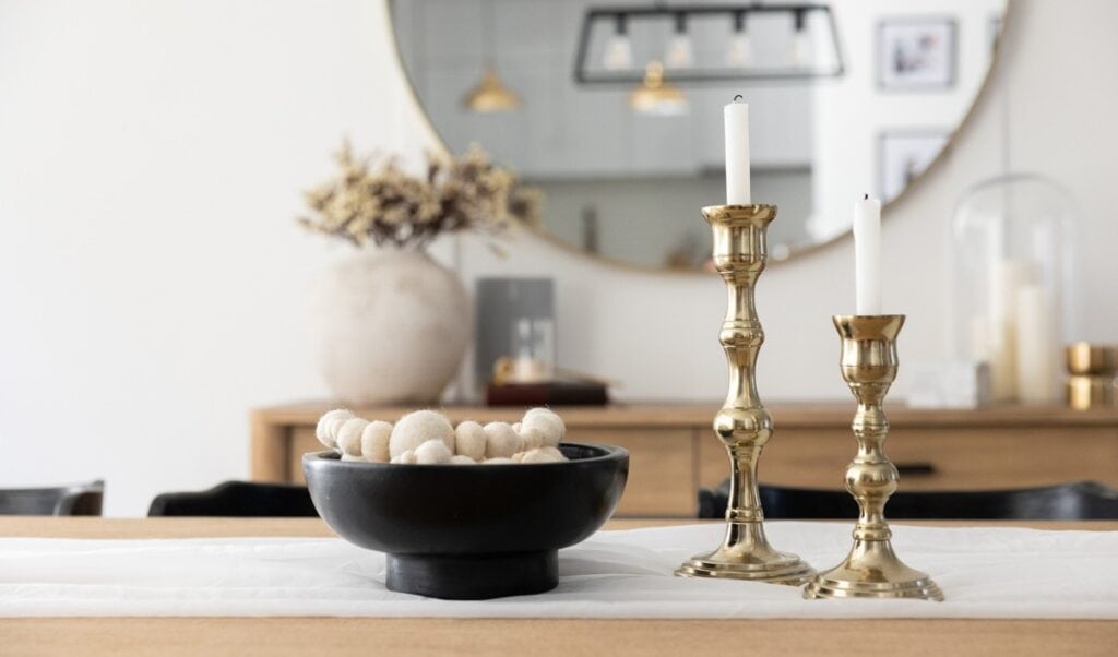 A modern dining table centerpiece featuring a black bowl filled with cream-colored felt balls sits beside two gold candlesticks with white taper candles. In the background, a large round mirror reflects pendant lighting and a neutral-toned room with minimalist decor.