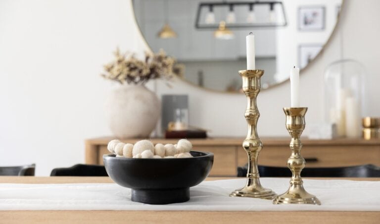 A modern dining table centerpiece featuring a black bowl filled with cream-colored felt balls sits beside two gold candlesticks with white taper candles. In the background, a large round mirror reflects pendant lighting and a neutral-toned room with minimalist decor.