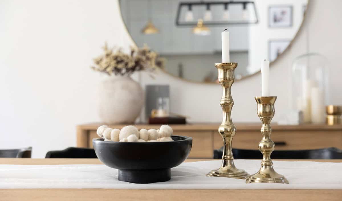 A modern dining table centerpiece featuring a black bowl filled with cream-colored felt balls sits beside two gold candlesticks with white taper candles. In the background, a large round mirror reflects pendant lighting and a neutral-toned room with minimalist decor.