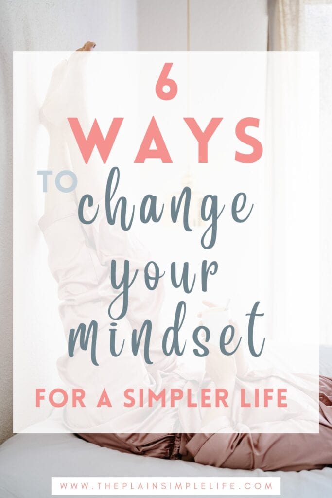 Pinterest pin with text overlay reading “6 ways to change your mindset for a simpler life.” A woman in pink loungewear relaxes with her legs resting up on a white wall while holding a mug, lying on a bed in a softly lit room with neutral decor.